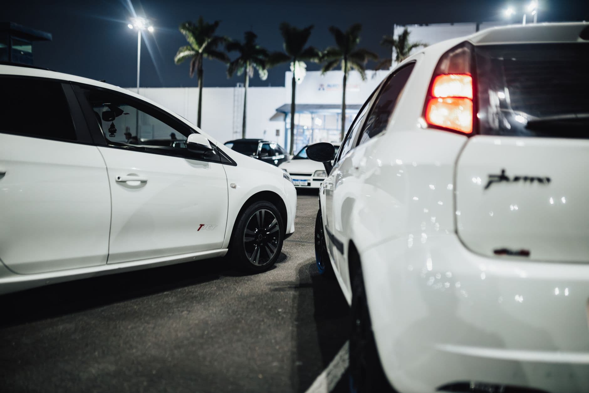 White cars gathered at a nighttime car meet in an illuminated parking lot.