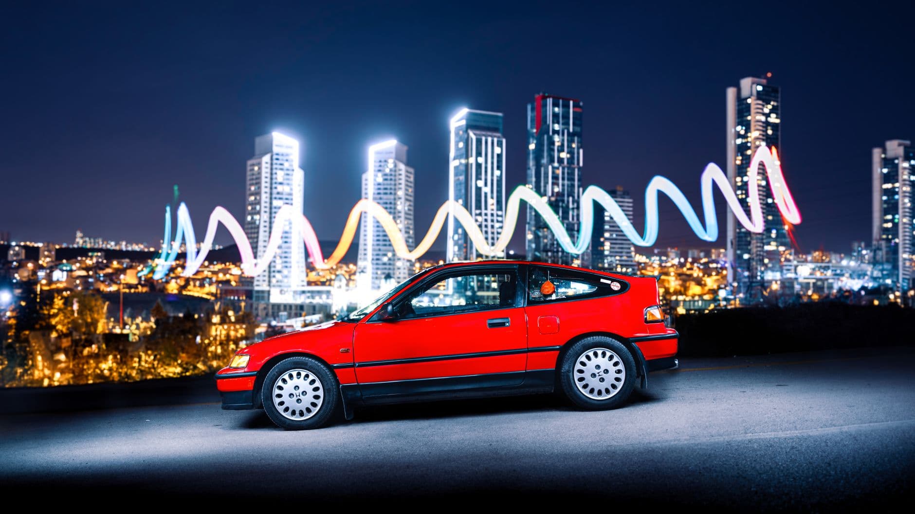 A vibrant red Honda CRX car with Ankara's night skyline as a backdrop, showcasing urban modernity.