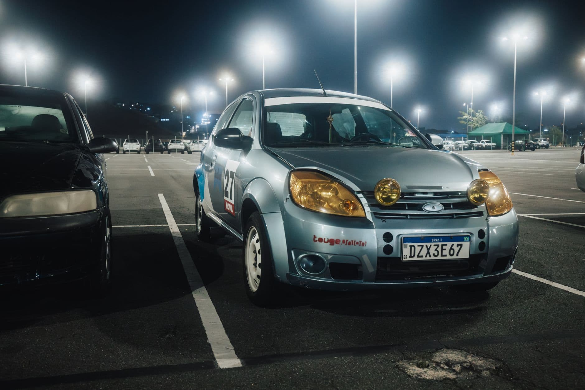Silver car with yellow headlights in a lit parking lot at night.