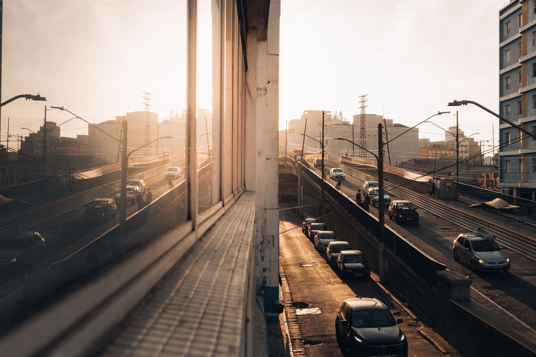 Cars drive on an urban highway during sunset, reflected in a building's windows, creating a warm atmosphere.